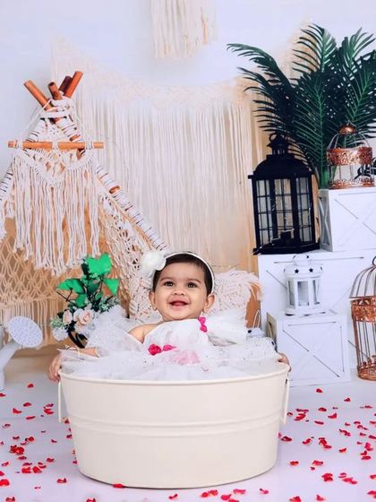 A happy baby girl enjoys a bubble bath in a white tub, surrounded by rose petals. The boho-chic background with a teepee and lanterns adds a stylish and whimsical touch.