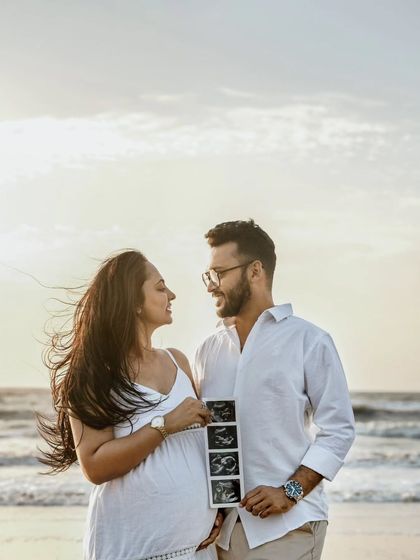 Announcing their joy at the beach. The couple is sharing a happy moment while holding up their baby's first pictures. The sunset and ocean waves make for a beautiful announcement setting.