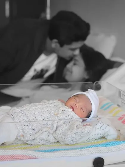 A tender moment with the newborn sleeping peacefully in the hospital bassinet while mom and dad share a loving kiss in the background. This shot tells a beautiful story of a new family beginning.