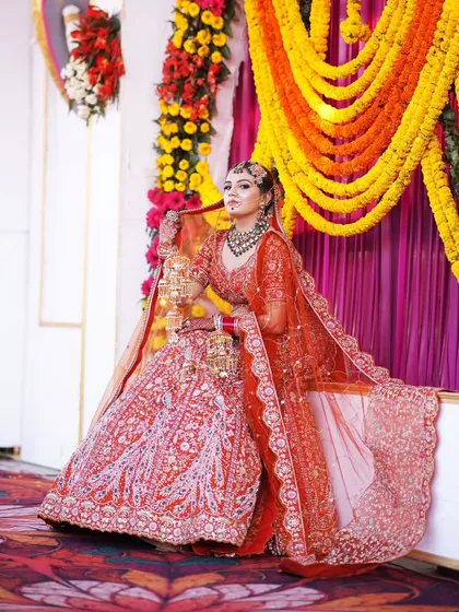 A full-length portrait of the bride seated, allowing her orange lehenga to spread out beautifully against the vibrant floral decor.