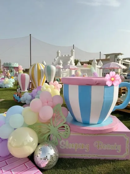 A whimsical outdoor setup featuring elements from Mickey Mouse's clubhouse, like a giant teacup prop and a bouncy castle in the background.