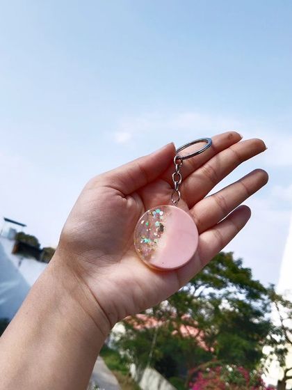 This circular keychain features a modern two-tone design with pastel pink and a clear section filled with holographic glitter. It's a chic and trendy accessory, shown here against a bright sky.
