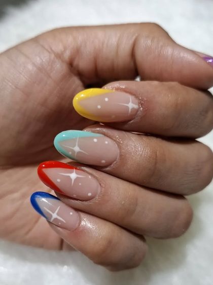 A playful rainbow French tip design. Each almond-shaped nail has a different colored tip, accented with tiny white dots and a sparkle for a fun, vibrant style.