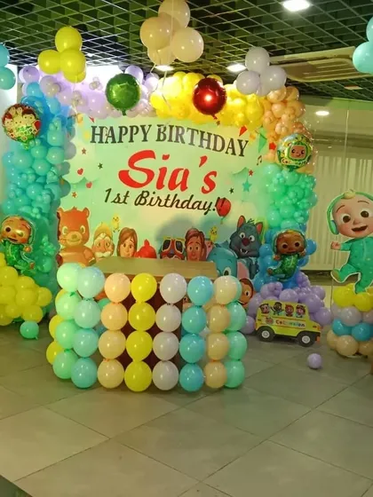 A complete Cocomelon first birthday stage setup for "Sia". The decoration includes a large themed backdrop, character cutouts, and a table made of balloons, all in bright, happy colors.