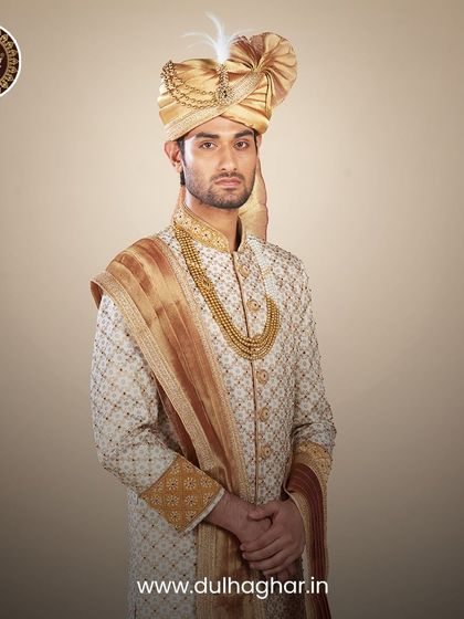 A full portrait of a groom in a cream and gold embroidered sherwani, complete with a safa and mala. This is a classic, unforgettable wedding day look.
