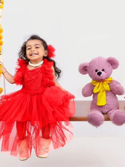 A happy girl in a red dress sits on a floral swing with her teddy bear. The simple white background and natural props create a clean and joyful portrait.