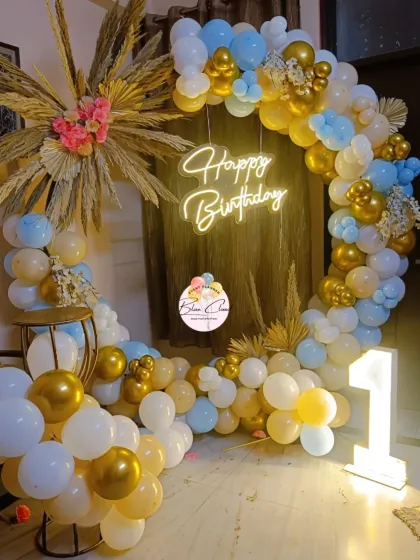 A charming and whimsical birthday setup with a boho-chic vibe. This decor features a circular balloon arch in pastel blue, white, and gold, accented with dried pampas grass and a 'Happy Birthday' neon sign.