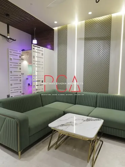 In the living room of 'The Cursive Abode' residence, comfort meets high design. The space is anchored by a custom green sectional sofa and a feature wall with textured panels and integrated vertical lighting, creating a warm and inviting atmosphere for family and guests.