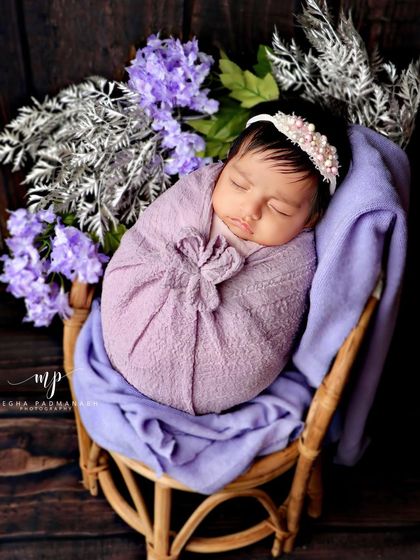 A baby wrapped in a purple flower-style swaddle, resting in a small rattan chair. The dark wood background makes the colors stand out.