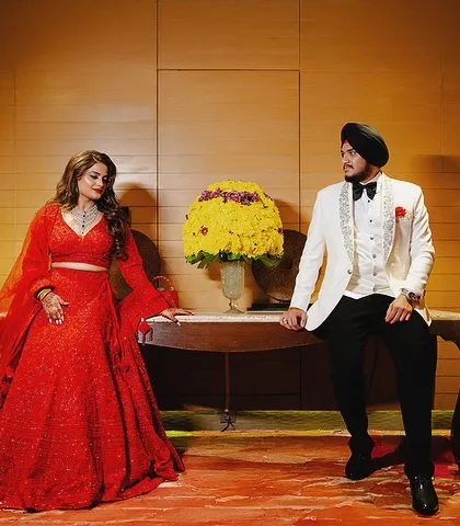 A full-length portrait showcasing the couple's stunning reception outfits. The bride's flowing red lehenga and the groom's white tuxedo make a bold statement.