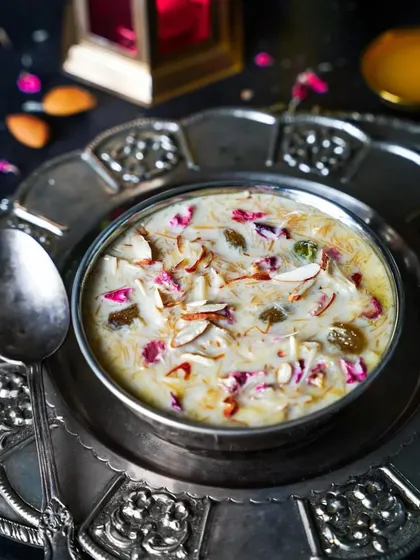 A luxurious bowl of Sheer Khurma, a traditional Eid dessert, garnished with nuts and rose petals. The dark, moody styling on a silver platter gives it a rich and festive feel.