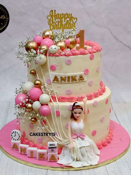 An 8kg chocolate showstopper, this two-tier cake combines a Barbie theme with festive balloons. Designed in pink and gold, it features a doll figure, a cascade of decorative balls, and personalized blocks.