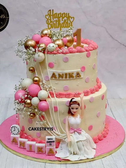 An 8kg chocolate showstopper, this two-tier cake combines a Barbie theme with festive balloons. Designed in pink and gold, it features a doll figure, a cascade of decorative balls, and personalized blocks.