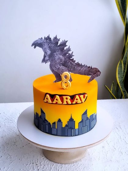 A Godzilla-themed cake for an eighth birthday. The cake features a city skyline silhouette against a fiery orange and yellow sunset, with a Godzilla topper.