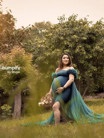 A wider shot of the expecting mother in the meadow, showcasing the full beauty of her gown against the natural landscape. The golden sun flare adds a magical glow to the scene.