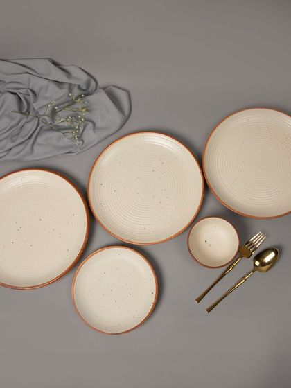 A flat-lay of a speckled, cream-colored dinnerware set with a rustic edge, styled with gold cutlery.
