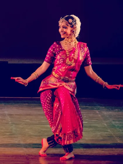 A vibrant stage photograph from a solo Bharatanatyam recital. The bright pink and gold costume, combined with the expressive smile, captures the joyful spirit of the dance.