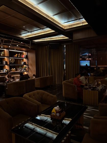 The interior of LimitlessLuxe in Noida showcases a moody and sophisticated ambiance. The dark wood tones, plush velvet seating, and strategic recessed lighting create an exclusive and comfortable lounge experience, perfect for a high-end dine-in spot.
