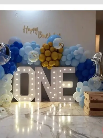 A stylish first birthday setup with large light-up 'ONE' letters as the centerpiece. The decoration is completed with a blue and gold balloon arrangement and a personalized bubble balloon.