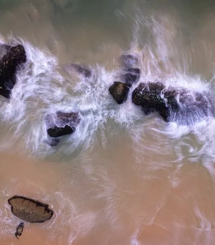 An abstract top-down long exposure of waves washing over rocks. The slow shutter speed creates a beautiful, swirling pattern of white water.