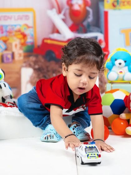 A toddler boy is deep in concentration as he plays with a toy car in a room filled with toys.