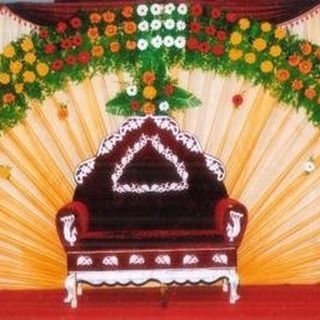 A simple stage setup with a fan-draped backdrop and a floral border. The focus is on the ornate wedding chair, making it suitable for a small function or ceremony.