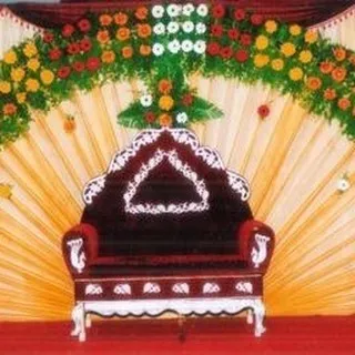 A simple stage setup with a fan-draped backdrop and a floral border. The focus is on the ornate wedding chair, making it suitable for a small function or ceremony.