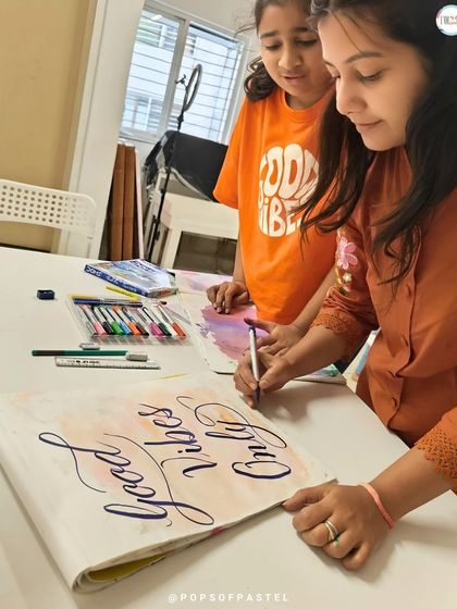 An instructor demonstrates how to form elegant, flowing letters for a "Good Vibes Only" piece, guiding a student through the process.