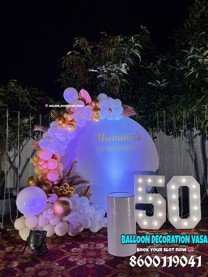 A 50th birthday celebration outdoors at night. The setup features a round backdrop with a balloon garland and large marquee numbers.