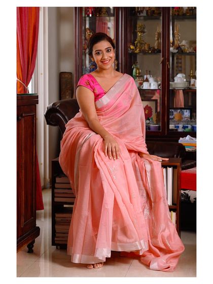 A relaxed and smiling portrait of a woman in a lovely pink saree, seated in a comfortable home environment.