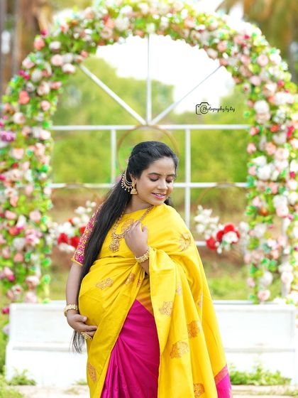 A beautiful solo portrait of a mother-to-be in a yellow and pink saree, framed by a floral arch.