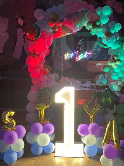 The party decoration glowing under red and green lights. The large marquee number '1' and the 'Happy Birthday' neon sign stand out, creating a vibrant party atmosphere.