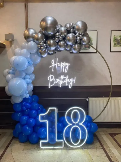 An 18th birthday setup with a half-ring balloon arch in shades of blue and silver, with neon signs for "Happy Birthday" and "18".