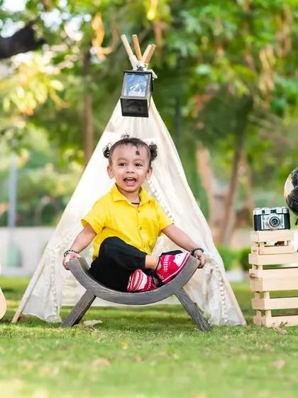 A one-year-old boy with a joyful expression sits in a traveler-themed outdoor setup for his pre-birthday photoshoot.