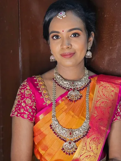 A classic muhurtham look with a bright orange and pink saree, complemented by silver jewelry with red stones. The makeup ties it all together beautifully.