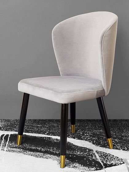 This elegant dining chair in a soft grey velvet provides plush seating and modern style. The legs feature a dark finish with gold-capped feet, adding a subtle touch of luxury and refinement.