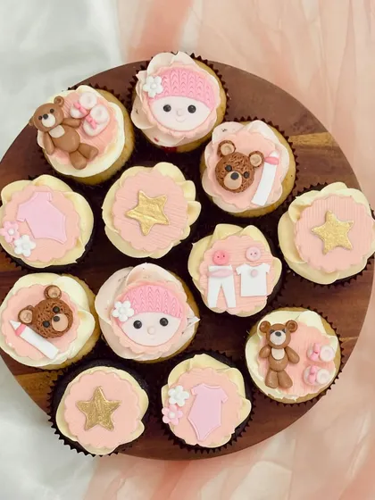 An assortment of pink-themed baby shower cupcakes on a wooden board. The designs include fondant teddy bears, baby clothes, gold stars, and baby faces with knitted hats, demonstrating a cohesive and detailed theme for celebrating a baby girl.