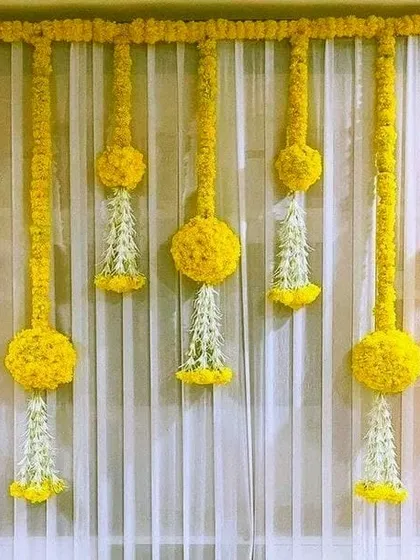 Simple details can make a big impact. For this Haldi background, we created an elegant look using hanging floral tassels made from marigold balls and white jasmine flowers against a sheer white curtain.