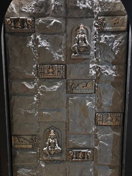 A feature wall for a mandir, constructed with rough-textured stone blocks and intricately carved deity figures. This creates a rustic yet divine atmosphere.
