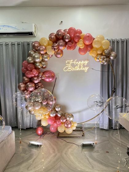 This is a duplicate of ID 17, 49, 92, 153, 209. An elegant balloon ring decoration for an intimate home celebration. This setup features a half-arch of chrome and pastel balloons on a golden ring, with a 'Happy Birthday' neon sign.