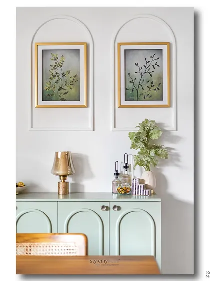 A mint green console with arched cabinet doors sits below two framed botanical prints. This symmetrical and calming composition showcases a love for gentle colors, clean lines, and nature-inspired art.