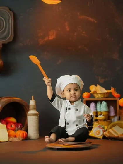 A creative and fun 'mini chef' themed photoshoot with a toddler dressed in a chef's uniform, playfully holding a wooden spoon in a rustic kitchen setup.
