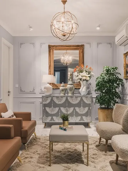 A beautifully composed shot of a neoclassical living area, focusing on the arrangement of the tan leather and plush white armchairs. The ornate chandelier and gilded mirror add a touch of classic luxury, while the artwork provides a splash of color.
