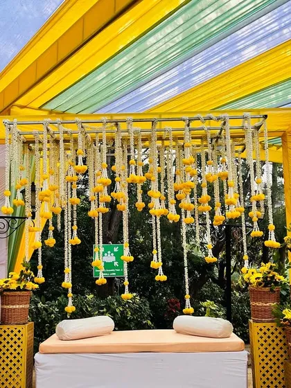 A bright and sunny Haldi stage setup with a canopy of yellow and light blue drapes. The stage is decorated with hanging floral tassels and baskets of sunflowers.