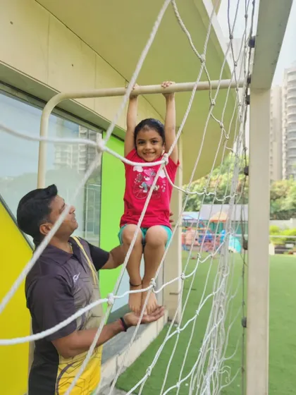 A smiling student enjoys her time on the climbing net, with a coach nearby to ensure her safety. We prioritize a supportive atmosphere where children can explore their physical abilities without fear.