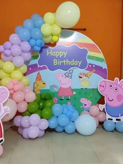 A playful Peppa Pig themed birthday decoration. This setup includes cutouts of the entire Peppa Pig family against a custom backdrop, framed by a pastel-colored balloon arch.