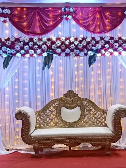 A classic wedding stage design featuring elegant red and white drapes. The backdrop is illuminated by a curtain of fairy lights, complemented by a floral cornice and matching bouquets for a timeless look.