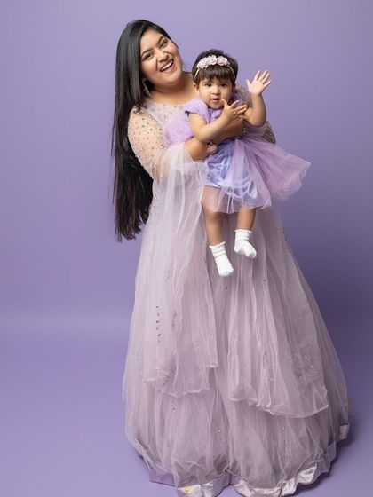 A mother and her daughter, dressed in matching lavender tones, smile brightly. The little one's happy wave is a delightful touch.
