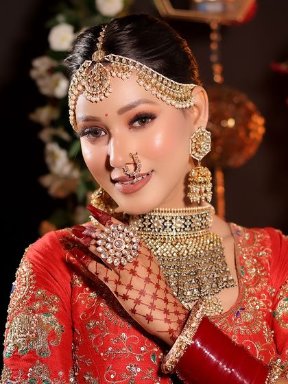 A smiling bride ready for her ceremony. The makeup features a soft glam eye, glowing cheeks, and a natural lip color, perfectly complementing her red lehenga and gold jewelry.