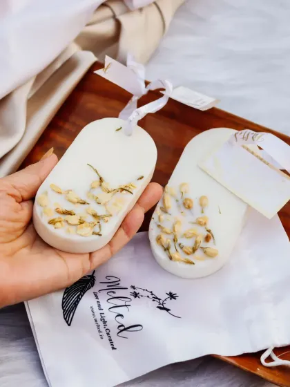 These beautifully handcrafted wax sachets, embedded with jasmine flowers, are held in hand to show their delicate detail. The soft lighting and natural wood tray create a warm, organic feel.
